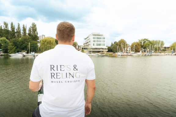 Mann mit Rückenansicht am Ufer eines Gewässers, trägt ein Shirt mit Aufschrift.