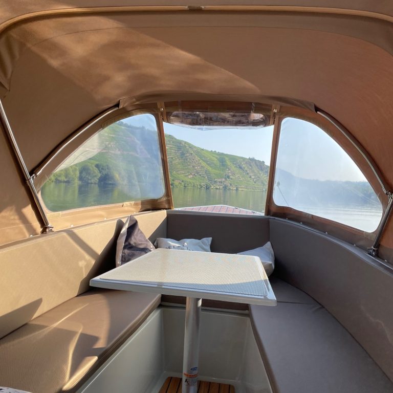 Innensicht eines Bootes mit Sitzgelegenheiten und Tisch, umgeben von grünen Hügeln.