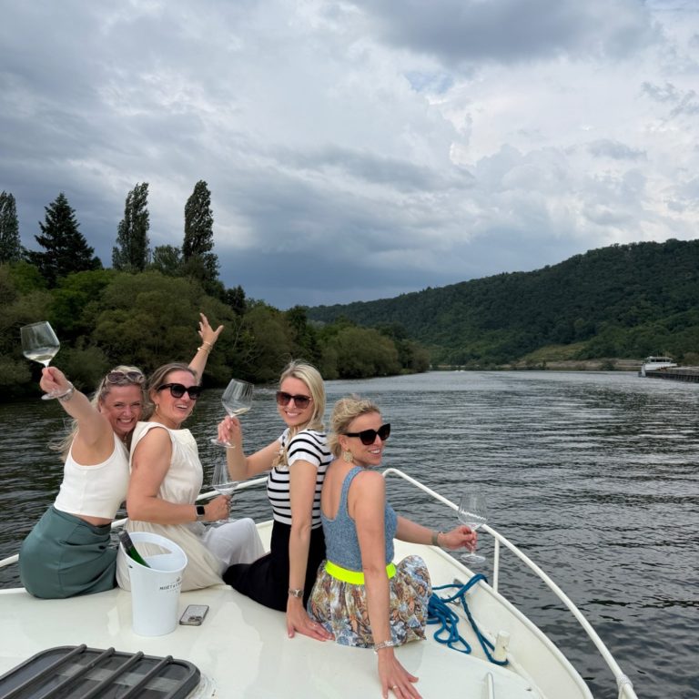 Vier Frauen auf einem Boot, genießen Drinks und die Aussicht auf einen Fluss und Hügel.