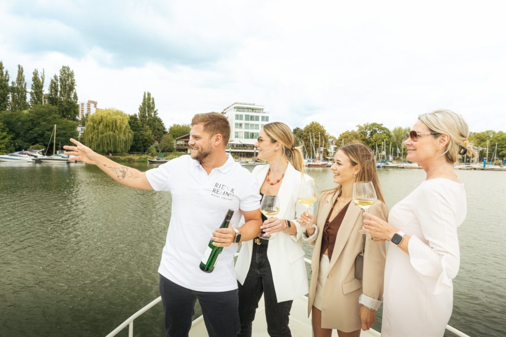 Vier Personen stehen auf einem Boot und schauen auf den See, einer zeigt mit dem Finger.