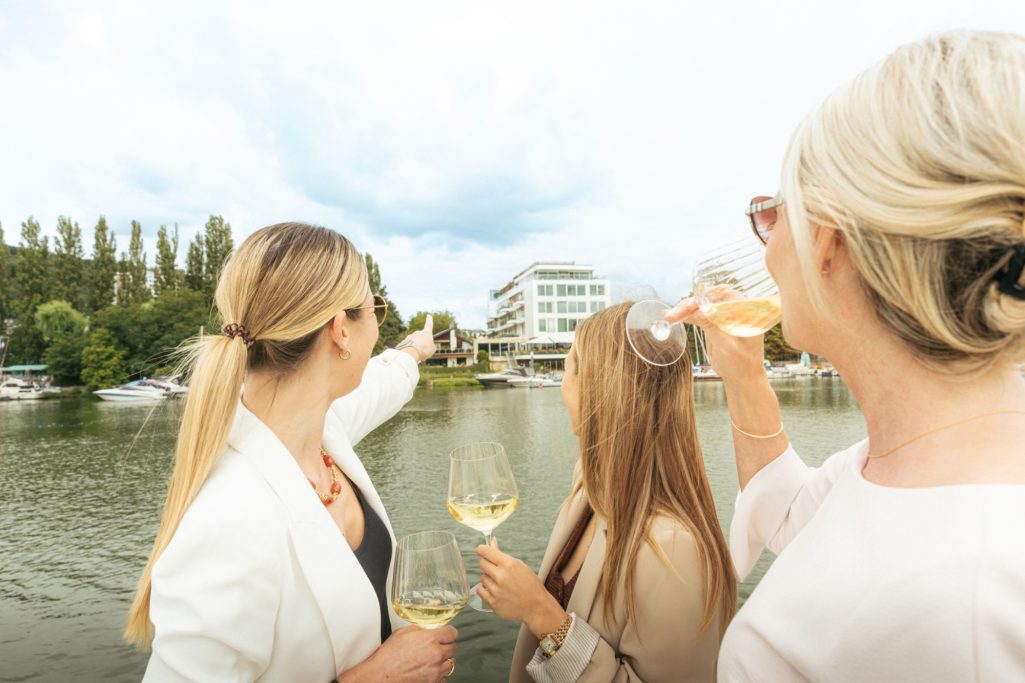Drei Frauen stoßen mit Gläsern am Wasser an und zeigen auf ein Gebäude am Ufer.