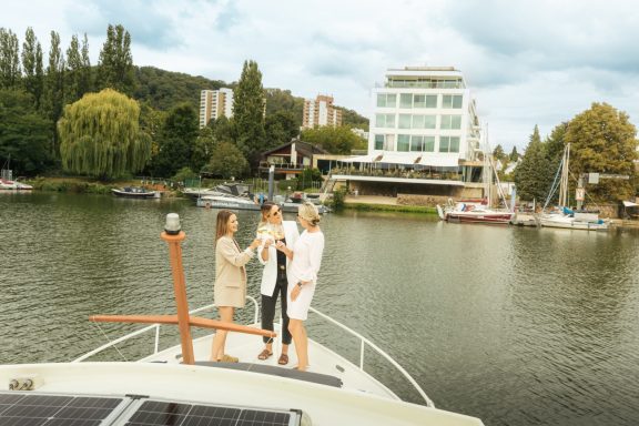 Drei Personen stehen auf einem Boot an einem ruhigen Gewässer mit einer modernen Gebäudeansicht.